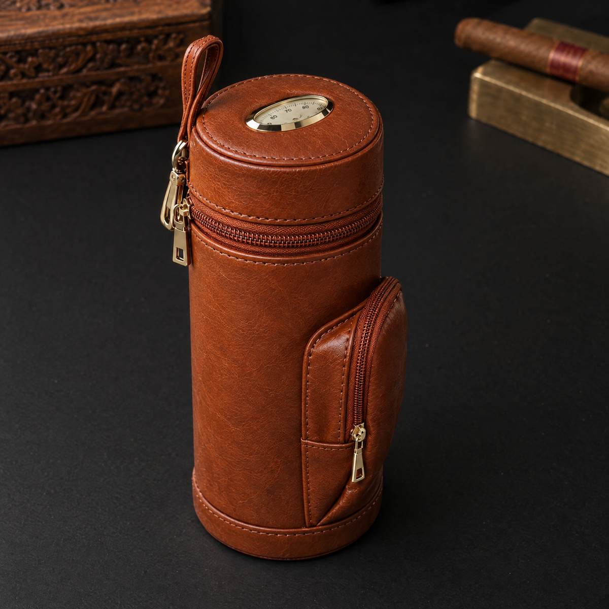 Cylindrical tan leather cigar travel case standing closed, with the hygrometer dial in the lid and a zipped front pocket